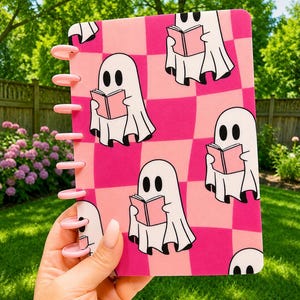 May include: A disc-bound notebook featuring a repeating pattern of white ghosts reading pink books against a background of alternating hot pink and light pink checkered squares. The notebook has light pink plastic binding discs along the left side.