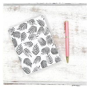 May include: A white notebook with a black and white leaf pattern design. A pink pen with a gold top is next to the notebook on a white wooden surface.