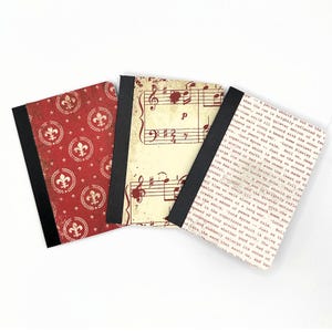 May include: Three small notebooks with black spines and different covers. One cover features a red and white pattern with a fleur-de-lis design. Another cover features a vintage sheet music design. The third cover features a text excerpt from a book.