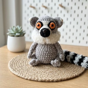 May include: A crocheted lemur plush toy featuring a gray textured body, a fluffy white neck ruff, bright orange eyes, and a black nose. The lemur has a long, plush tail with alternating black and white stripes, sitting in an upright position.