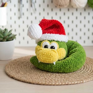 Puede incluir: Un juguete de serpiente de ganchillo en verde y amarillo que lleva un gorro de Papá Noel rojo y blanco con un pompón blanco. La serpiente tiene grandes ojos blancos con pupilas negras y dos pequeños colmillos blancos en su hocico amarillo.