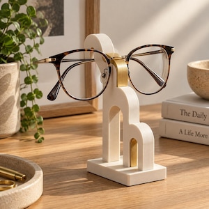 May include: A white marble eyeglass stand with a gold metal bridge, holding a pair of tortoise shell glasses. The stand features a tiered arch design on a rectangular base, providing a stable structure for displaying eyewear on a desk.