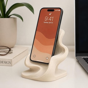 May include: A light-colored, wavy, layered wooden phone stand holding a black smartphone with a screen displaying the time 9:41 and the date Tuesday, April 23. The stand features a smooth, organic, tiered design that supports the device upright.