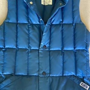 May include: A blue sleeveless puffer vest featuring a high collar, a front button closure, and vertical quilted stitching. The garment includes two front pockets and a white label with the text PUFFA on the lower left pocket and inside the collar.