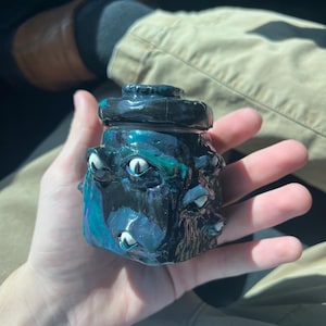 May include: A small, dark, glossy jar with a lid, featuring multiple sculpted eyes embedded into the surface. The jar has a swirling colour palette of deep blue, black, and hints of purple, with white and black iris details on the protruding eyes.