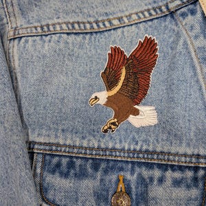 Può includere: Una giacca di jeans azzurro lavaggio acido con una toppa ricamata di un'aquila calva in volo. L'aquila ha ali marroni e bruno-rossastre, testa bianca e coda bianca. Un bottone in metallo è visibile sulla patta della tasca sul petto.