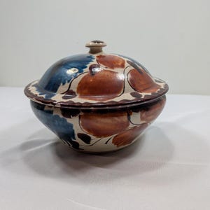 May include: A round ceramic lidded bowl featuring a cream-colored base with hand-painted floral motifs in shades of rust-brown and deep blue. The lid has a small central knob and a wide rim, matching the bowl's decorative pattern and glossy finish.