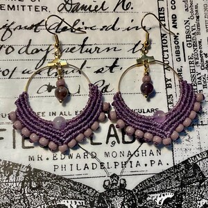 May include: A pair of handmade hoop earrings. The earrings feature a gold-colored hoop with a woven purple macrame design. The macrame is adorned with small, light purple beads and larger, round purple beads.