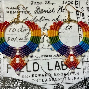 May include: Rainbow macrame hoop earrings with gold-colored hooks. The earrings feature a vibrant spectrum of colors, including red, orange, yellow, green, blue, and purple. White beads are attached to the bottom of the earrings. The earrings are on a vintage paper background.