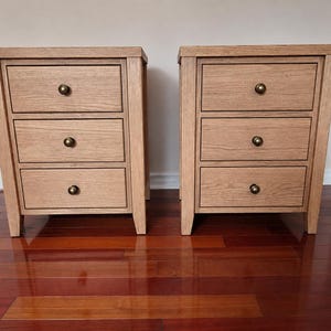 May include: Two matching light brown wooden bedside tables, each featuring three stacked drawers with visible wood grain patterns. Every drawer is equipped with a single small, round, dark metallic knob centred on the front panel.