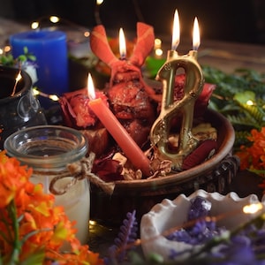 May include: A decorative arrangement featuring a gold-colored number eight candle, a slender pink taper candle, and a red heart-shaped object, all lit and placed within a dark bowl. The scene includes orange flowers, a glass jar, and purple crystals.