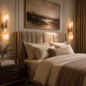 May include: Two wall-mounted light fixtures featuring dual vertical fluted glass shades with a gold-toned metal base. Each fixture emits a warm glow, illuminating the wall on either side of a beige upholstered headboard in a bedroom setting.