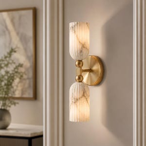 May include: A wall sconce featuring two vertically stacked, fluted cylindrical shades made of white marble with grey veining. The shades are connected by a central brass-toned metal rod and mounted to the wall with a circular brass-toned backplate.
