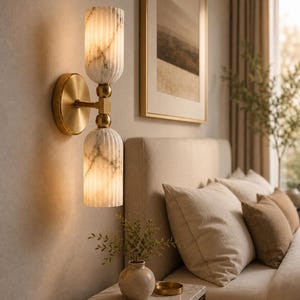 May include: A wall-mounted light fixture featuring a circular brass base and a central brass arm connecting two vertically stacked, cylindrical, fluted white marble glass shades that emit a warm, soft glow from within the textured glass.