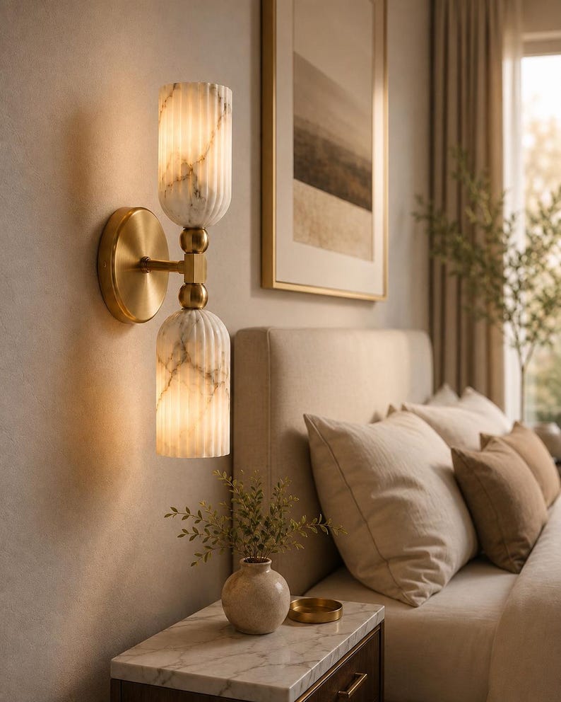 May include: A wall-mounted light fixture featuring a circular brass base and a central brass arm connecting two vertically stacked, cylindrical, fluted white marble glass shades that emit a warm, soft glow from within the textured glass.