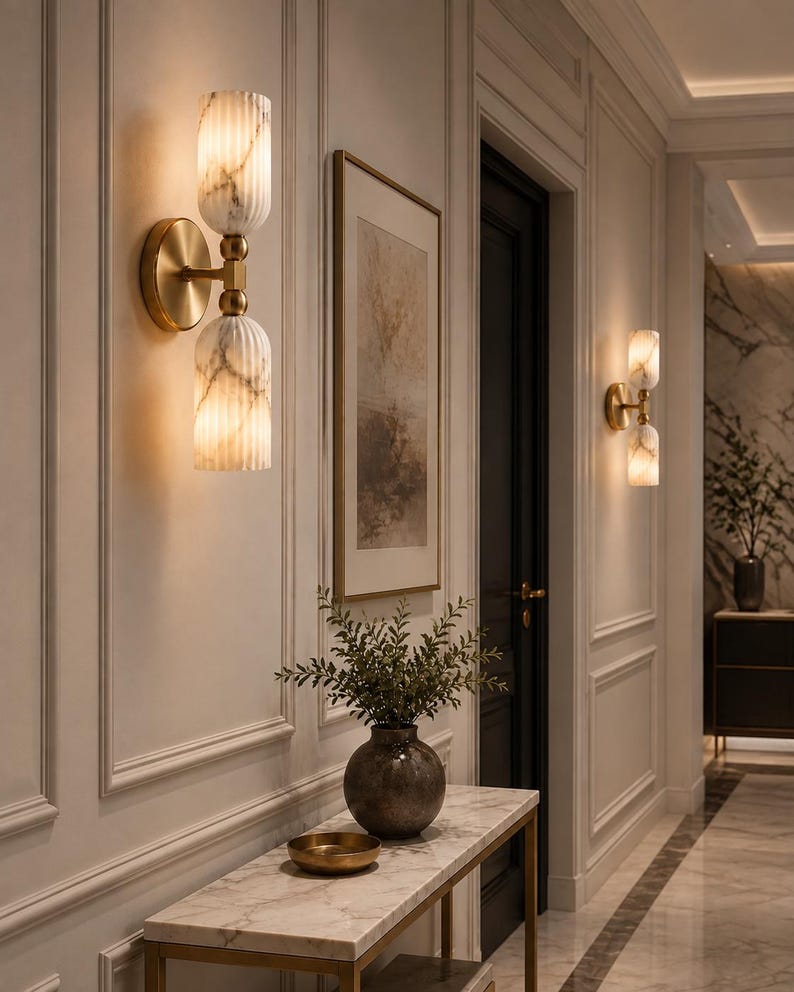 May include: A wall sconce featuring two vertically stacked, fluted, white marble-patterned glass cylinders connected by a brass-toned metal fixture. The light fixture emits a warm glow from both the top and bottom glass shades mounted on a wall.