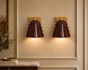May include: Two wall sconces featuring deep burgundy, bell-shaped shades with vertical ridges. Each shade is accented by three small, spherical brass-toned studs on each side and mounted on a square brass-toned backplate, emitting a warm light.
