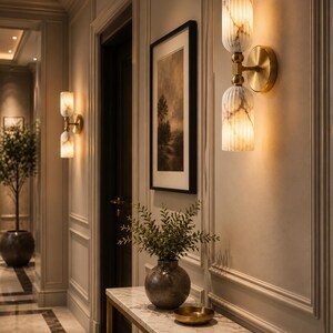 May include: A wall-mounted light fixture featuring two vertically stacked, fluted, white marble-patterned glass shades connected by a central brass-toned metal arm and a circular wall plate, emitting a warm, soft glow against a paneled wall.