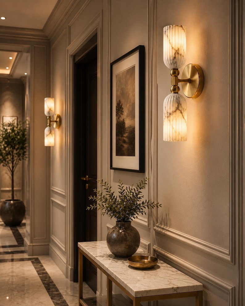 May include: A wall-mounted light fixture featuring two vertically stacked, fluted, white marble-patterned glass shades connected by a central brass-toned metal arm and a circular wall plate, emitting a warm, soft glow against a paneled wall.