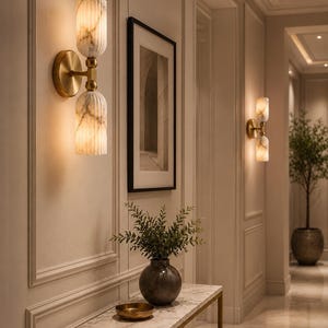 May include: A wall sconce featuring two vertically stacked, fluted, white marble-patterned glass shades connected by a brushed brass central fixture and a circular wall-mounted base, emitting a warm, soft glow from within the glass cylinders.