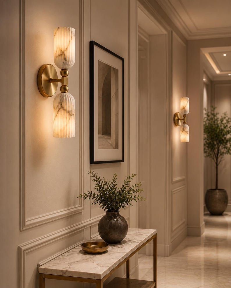 May include: A wall sconce featuring two vertically stacked, fluted, white marble-patterned glass shades connected by a brushed brass central fixture and a circular wall-mounted base, emitting a warm, soft glow from within the glass cylinders.