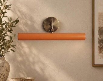 May include: A wall-mounted picture light featuring a long, cylindrical orange shade attached to a circular, antique brass-finished metal base. The fixture has a central arm connecting the horizontal tube to the wall plate, providing a sleek design.