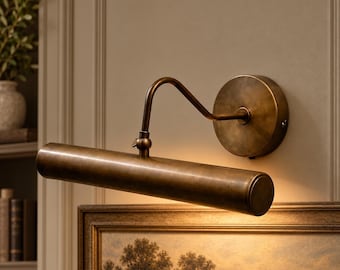 May include: A wall-mounted picture light with an antique brass finish. The fixture features a circular base plate, a curved arm, and a long, cylindrical horizontal lamp head that casts a warm downward glow over a framed landscape painting.