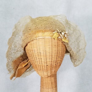 May include: A vintage beige lace hat with a gold satin ribbon and a floral embellishment. The hat is displayed on a wicker hat stand.