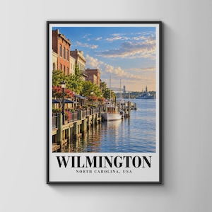 May include: A framed vertical wall art print depicting the Wilmington, North Carolina waterfront. The scene features historic brick buildings along a wooden pier, boats docked in the water, and a large ship in the distance under a blue sky.