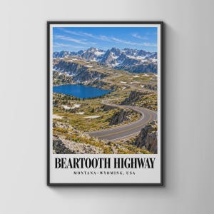 Op de afbeelding: Een ingelijste verticale wandkunstprint met een kronkelende asfaltweg door een berglandschap met een diepblauw meer en besneeuwde bergtoppen onder een heldere lucht. De tekst onderaan luidt BEARTOOTH HIGHWAY MONTANA-WYOMING, USA.