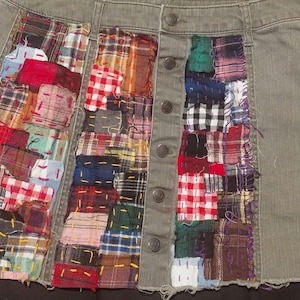 May include: An olive green denim skirt featuring a button-front closure and two vertical panels of multicolored patchwork fabric. The patchwork sections consist of various plaid and checkered patterns in red, blue, yellow, white, and black fabric.