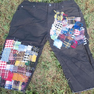 May include: Black denim shorts featuring extensive patchwork detailing on the front panels. The patches consist of various colorful plaid, checkered, and solid fabric squares stitched onto the legs with visible, contrasting thread work.