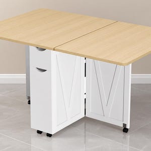 Pode incluir: Uma mesa dobr&aacute;vel com um tampo retangular de madeira clara e uma base branca. A base possui duas unidades de armazenamento verticais com puxadores embutidos e pain&eacute;is laterais com um design em V. Toda a estrutura &eacute; apoiada por pequenas rodas pretas.