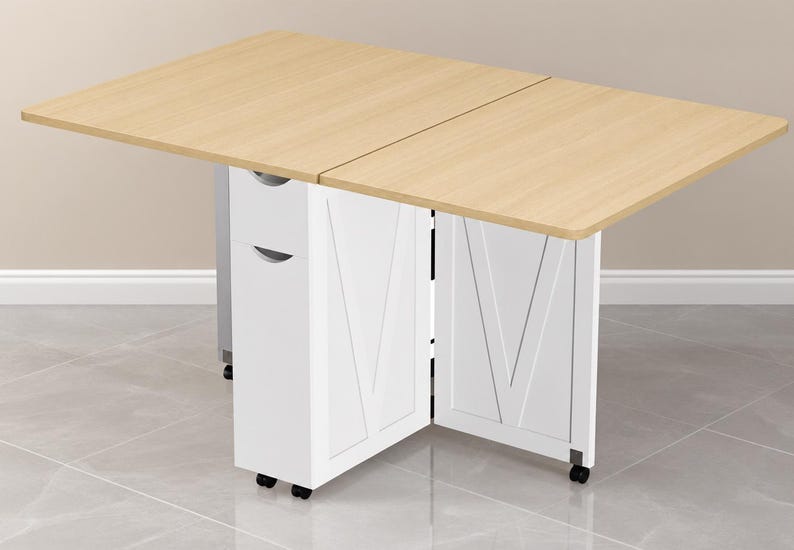 Pode incluir: Uma mesa dobr&aacute;vel com um tampo retangular de madeira clara e uma base branca. A base possui duas unidades de armazenamento verticais com puxadores embutidos e pain&eacute;is laterais com um design em V. Toda a estrutura &eacute; apoiada por pequenas rodas pretas.