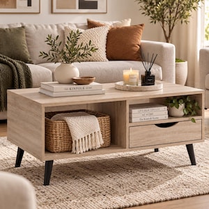 May include: A light wood coffee table with four black tapered legs, featuring an open shelf containing a woven basket with a white fringed throw, a single drawer, and a side compartment holding books titled Live Beautifully At Home and The Kinfolk Home.