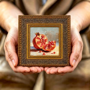 May include: A small square oil painting in an ornate, dark bronze-coloured frame. The artwork depicts a halved pomegranate with vibrant red seeds exposed, resting on a light-coloured surface against a soft, muted, neutral-toned background.