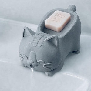 Pode incluir: Uma saboneteira cinza fosca em formato de gato com superfície texturizada e uma pequena cauda. O gato está posicionado com a boca aberta, permitindo que a água escorra. Um sabonete retangular rosa claro com o texto "SPICE ECHCE" está em cima.