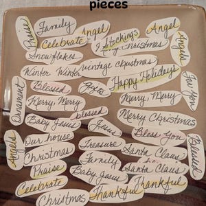 May include: A set of 39 paper cutouts labeled Holiday Word Mix, each featuring handwritten holiday-themed words like Christmas, Santa Claus, and Blessed in black ink on white oval-shaped paper pieces, arranged on a square brown plate.