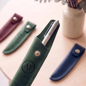 Puede incluir: Una funda de cuero verde bosque para bolígrafo con un botón de presión de metal plateado y las letras AF grabadas dentro de un borde circular. Un bolígrafo plateado está insertado en la funda. Se ven otras fundas en burdeos, verde y azul marino.