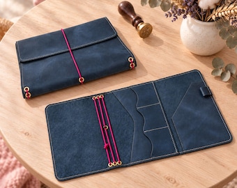 May include: Two dark blue leather notebook covers with a matte finish. One cover is folded shut with a bright pink elastic band, while the other is open to reveal internal pockets, slots, and three vertical pink elastic cords with gold eyelets.