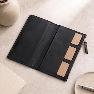 Puede incluir: Una cartera de cuero negra de forma rectangular alargada, con un cierre de cremallera de metal dorado y una lengüeta de cuero. El interior incluye tres ranuras verticales para tarjetas y un compartimento abierto para billetes o notas.