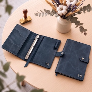May include: Two dark blue leather ring binders with a smooth, matte finish. One binder is open, revealing a silver three-ring mechanism and interior pockets. The other binder is closed with a snap button closure. Both feature the gold initials VB.