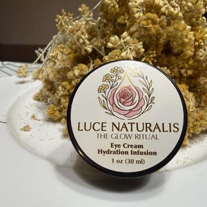 Op de afbeelding: Een ronde pot Luce Naturalis The Glow Ritual Eye Cream Hydration Infusion, 30 ml. Het etiket bevat een roze roosillustratie met gouden accenten, zwarte tekst en een witte achtergrond, geplaatst voor gedroogde gele bloemen.