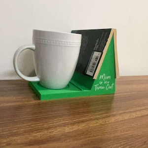 May include: A white ceramic mug with a textured rim sits on a green wooden stand shaped like a house. The stand features the white text "Mom is in a Time Out" and holds a black book upright against its angled back support.
