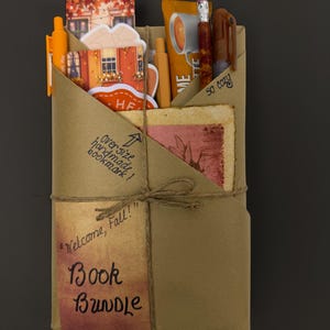May include: A brown paper wrapped book bundle tied with twine, featuring handwritten text "Welcome, Fall!" and "Book Bundle". An oversized handmade bookmark, orange pens, a pencil, and a coffee packet are tucked into the top folds of the paper.