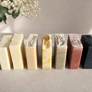 Könnte beinhalten: Eine Reihe von sieben rechteckigen Seifenstücken in verschiedenen Farben und Texturen, darunter einfarbiges Weiß, Creme, Beige, ein gelb gesprenkeltes Stück mit getrockneten Blüten, Grau, Altrosa und einfarbiges Schwarz, horizontal nebeneinander.