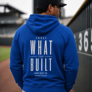 May include: A royal blue hooded sweatshirt featuring white text on the back that reads TRUST WHAT YOU BUILT, ZERO HYPE CO. BASEBALL DIVISION. The hoodie has long sleeves, a drawstring hood, and a ribbed hem, worn with light-coloured trousers.