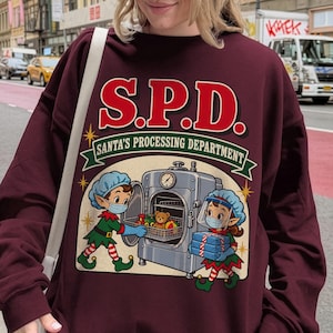 May include: A maroon long-sleeve sweatshirt featuring a graphic print with the text S.P.D. SANTA'S PROCESSING DEPARTMENT. The design shows two elves in hairnets and face masks working with a large metal machine containing toys and gift boxes.
