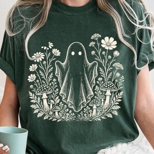 May include: A dark green short-sleeve t-shirt featuring a cream-colored graphic print of a central ghost figure surrounded by various wildflowers and mushrooms. The shirt has a crew neckline and a relaxed fit, showcasing a detailed botanical illustration.