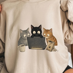 Op de afbeelding: Een beige sweater met ronde hals voorzien van een grafische illustratie van drie katten rondom een laptop. Een grijze kat zit naast een koffiemok, een zwarte kat typt op de laptop en een oranje cyperse kat zit naast een papieren zak en een muis.
